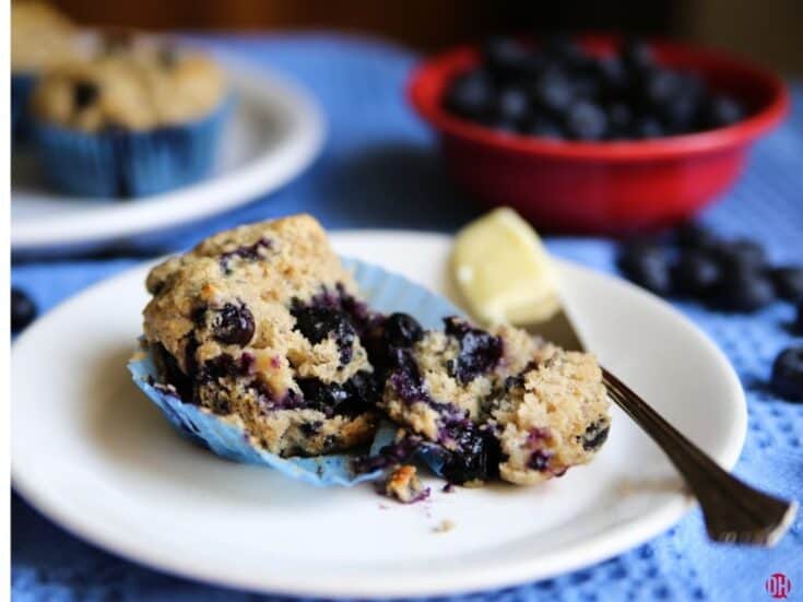 blueberry muffin halfed on white plate with knife with butter on it.